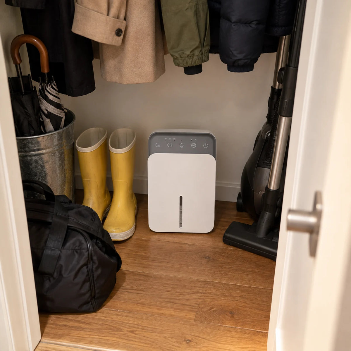 Mini déshumidificateur au fond d'un placard d'entrée, entre des bottes jaunes et un aspirateur